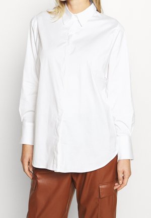 Femme portant une chemise blanche à manches longues boutonnée et un pantalon en cuir marron avec des poches latérales, debout devant un fond uni.