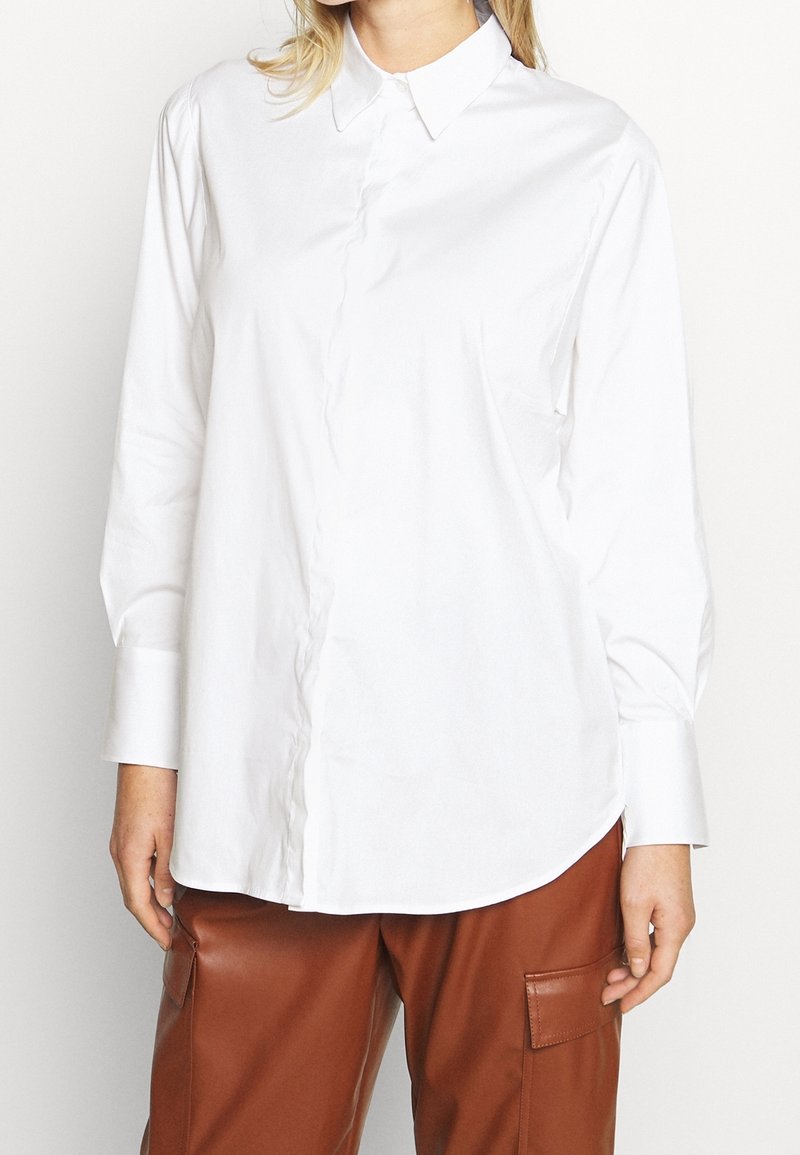 Femme portant une chemise blanche à manches longues boutonnée et un pantalon en cuir marron avec des poches latérales, debout devant un fond uni.