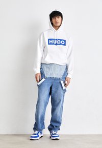 Hvit hettegenser med blå "HUGO"-logo, kombinert med lyseblå overaller med metall hardware og avtakbare detaljer; fullført med blå og hvite sneakers.