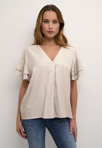 Beige V nyakú blúz, réteges fodros ujjakkal, sima anyagból készült. Világoskék farmerral viselve, laza szabással és oldalsó hasítékokkal.