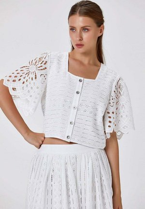 Femme portant un crop top en dentelle blanc boutonné avec des manches à découpes florales et une jupe en dentelle taille haute assortie sur un fond uni.