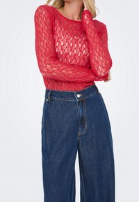 Femme portant un haut en dentelle rouge à manches longues et un jean large en denim bleu foncé taille haute, un bras croisé et l'autre main près du cou.