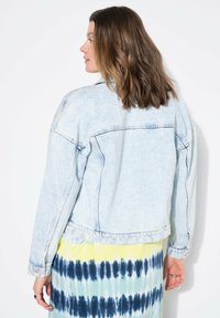 Hellblaues Denim-Jackett mit langen Ärmeln, lässiger Passform und genähten Taschen. Der Stoff hat ein verblasstes Finish und wird mit einem mehrfarbigen Batik-Kleid kombiniert.