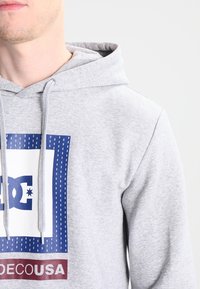 Grå huvtröja med en känguruficka, med en blå och vit logotypdesign och röda detaljer längst ner. Bomullstextur, avslappnad passform.