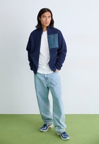 Fleece jacket in navy with a teal chest pocket, paired with light blue loose jeans and blue sneakers, set against a light background.