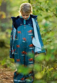 Blå vandtæt overall med købmønster, lange ærmer og en frontlynlås, båret over en mørkeblå top, sat i et naturligt udendørs miljø.