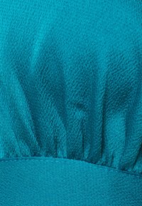 Tecido teal com uma superfície texturizada. Apresenta detalhes franzidos ao longo da borda superior, criando um efeito de volantes. Seção inferior lisa.