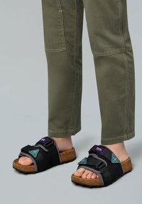 Sandali neri con suole in sughero, dotati di cinghie regolabili e dettagli in teal, indossati con pantaloni verde oliva, che mettono in risalto un design casual.