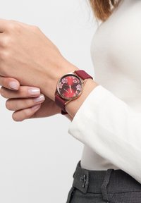 Runde Uhr mit Zifferblatt in rotem Blumenmuster, goldenen Akzenten und einem mahagoni-farbenen Silikonarmband. Getragen an einer Hand vor einem hellen Hintergrund.