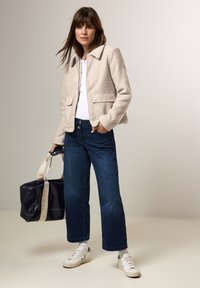 Beige textuur jasje met een kraag, twee zijzakken, gecombineerd met donkere denim wijde pijpen jeans en witte sneakers. Zwarte en beige tas.