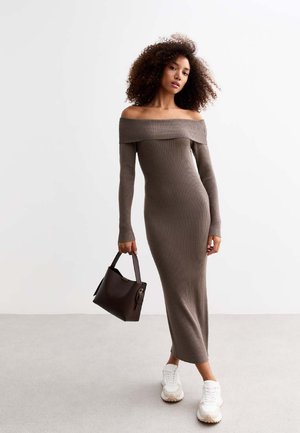 Robe longue à bretelles dénudées en rib, de couleur marron, au design ajusté. Le modèle tient un sac à main marron foncé et porte des baskets blanches. Texture lisse.