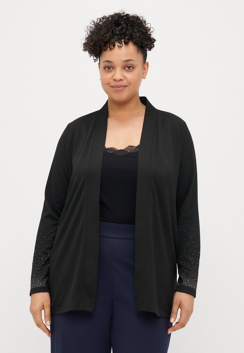 Μαύρο cardigan με ανοιχτό μπροστινό μέρος, υφασμάτινη υφή, μακριά μανίκια διακοσμημένα με μια κλίμακα από μικρές τρουκς, φοριέται πάνω από μια μαύρη μπλούζα.