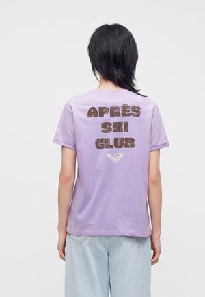 Persoană cu păr negru scurt, purtând un tricou lila deschis cu textul "APRÈS SKI CLUB" pe spate și blugi albastru deschis.