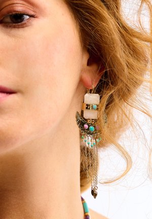 Hängende Ohrringe aus verschiedenen Materialien: weißer Stein, goldene Akzente und bunte Perlen. Ketten verleihen mit filigranen Details zusätzliche Struktur.