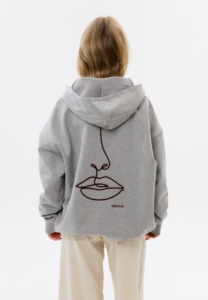 Persona con capelli biondi che indossa una felpa grigia con cappuccio, con un'illustrazione astratta del volto a linee e la scritta "MIRONS" sul retro, abbinata a pantaloni beige chiaro.