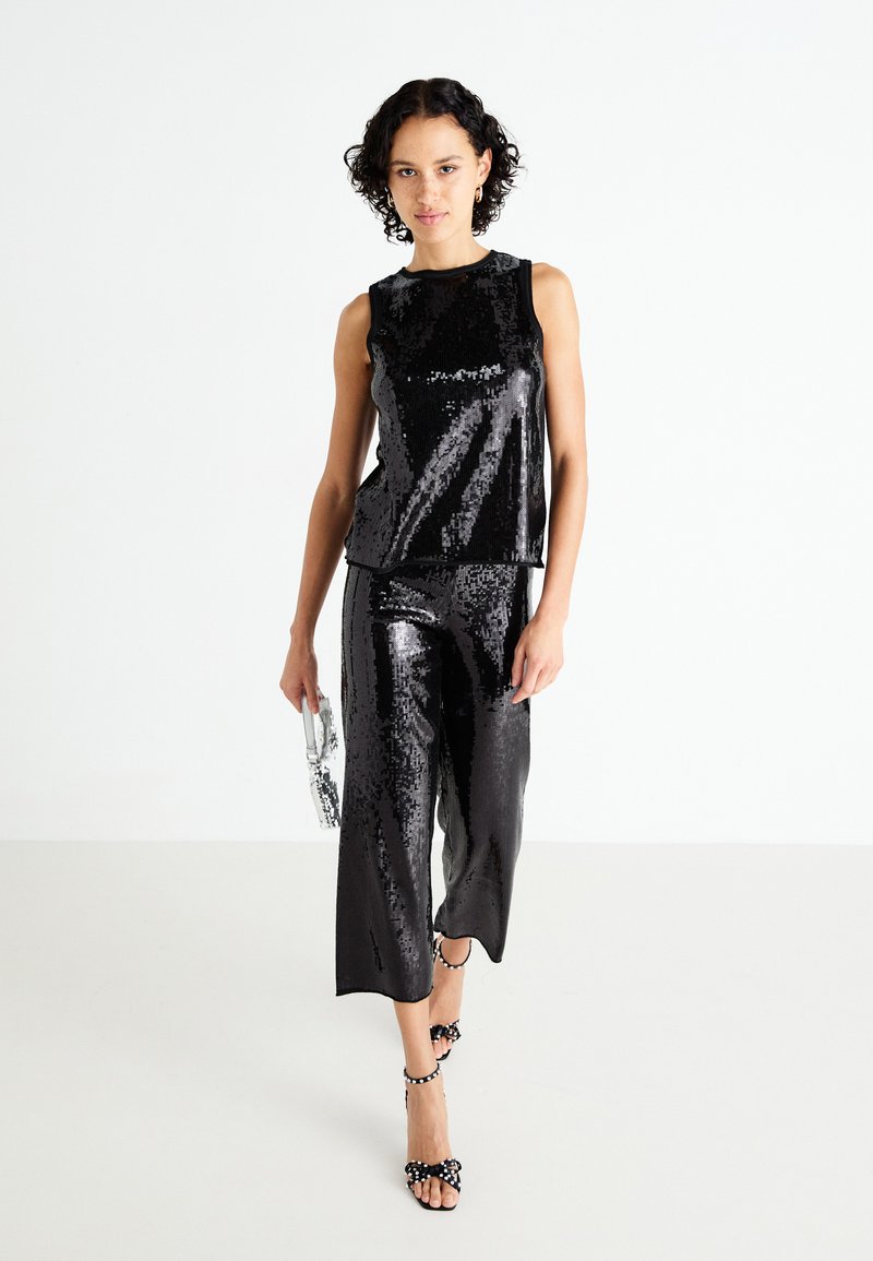Schwarzes Paillettentop und passende, kurz geschnittene Hose mit glänzendem Finish. Das Model hält eine durchsichtige Tasche und trägt schwarze High Heels mit einer Schleifendetails.