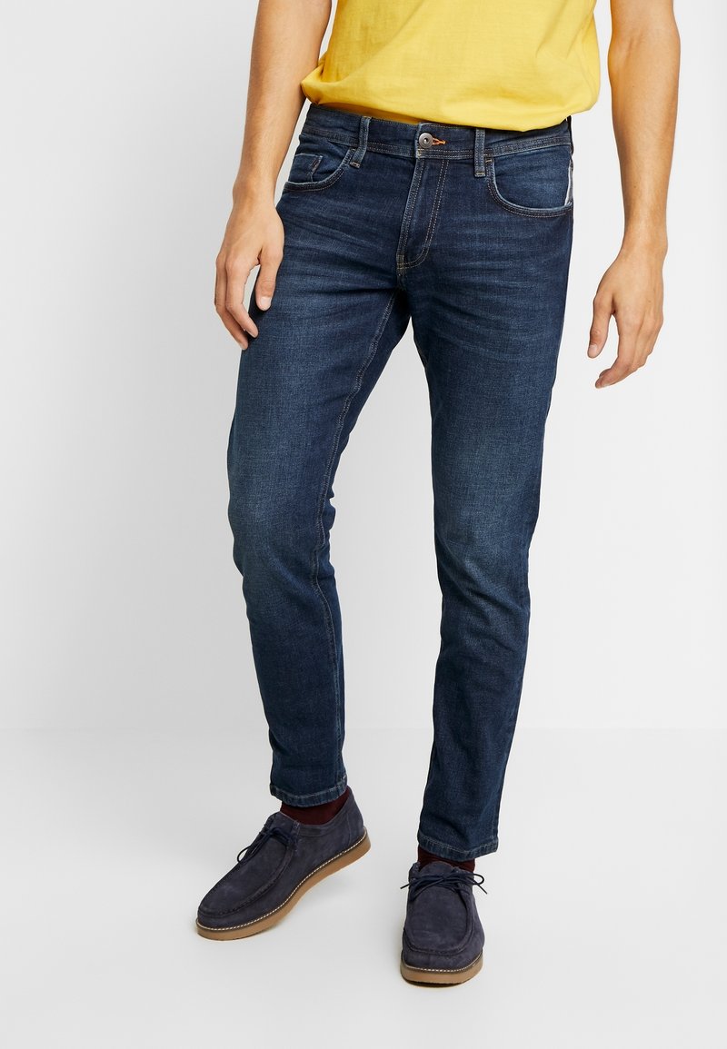 Homme portant un jean slim bleu foncé, une chemise jaune, des chaussettes bordeaux et des chaussures à lacets bleu marine, debout sur un sol blanc.