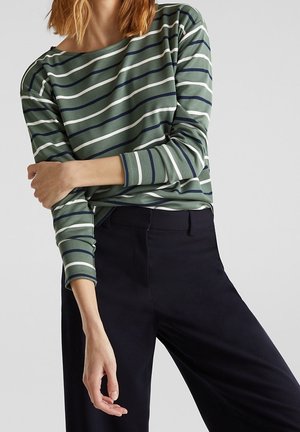 Osoba ubrana w zielono-białą pasiastą koszulę z długim rękawem i granatowe spodnie, z założonymi rękami, jedną ręką dotyka przeciwległego łokcia.