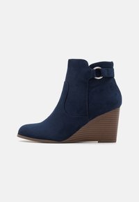 Bottes à talons compensés bleu marine avec une texture en suède, bout arrondi, élastique latéral et une sangle décorative avec un accent métallique circulaire.