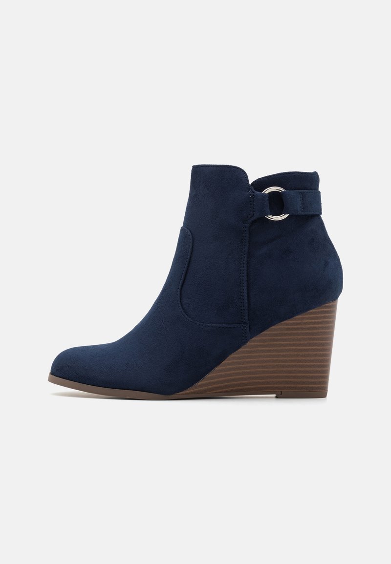 Bottes à talons compensés bleu marine avec une texture en suède, bout arrondi, élastique latéral et une sangle décorative avec un accent métallique circulaire.