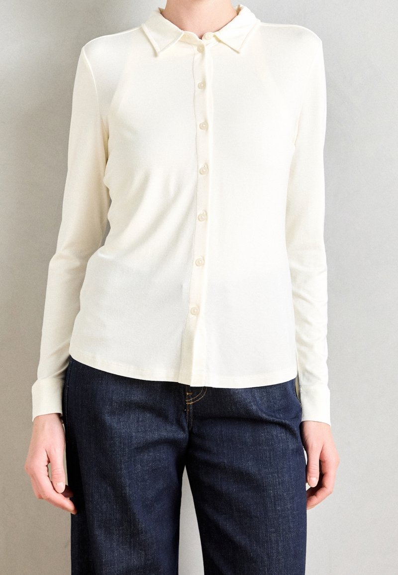 Personne portant une chemise blanche à manches longues avec col et boutons, et un jean bleu foncé, sur un fond clair uni.