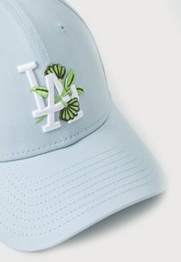 Ljusblå baseballkeps i tyg, med en vit broderad "LA"-logotyp som har gröna blad och fjärils detaljer, strukturerad design.