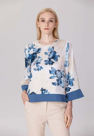 Femme blonde portant un chemisier blanc à motif floral bleu et larges poignets, associée à un pantalon beige, debout devant un fond uni.