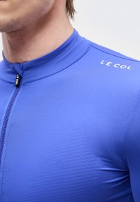 Gros plan d'une personne portant un maillot de cyclisme bleu avec une fermeture éclair à l'avant et le logo "LE COL" sur l'épaule et le tire-zip.