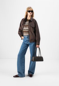 Femeie purtând ochelari de soare negri, jachetă maro, pulover cu model, blugi largi albaștri, ținând o geantă maro închis, stând în fața unui fundal alb.