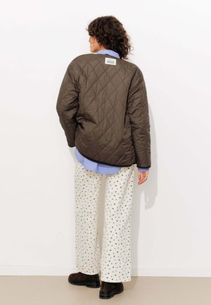 Veste matelassée marron avec un motif en losanges, portée sur une chemise bleu clair. Associée à un pantalon crème à motifs et des bottes marron foncé.