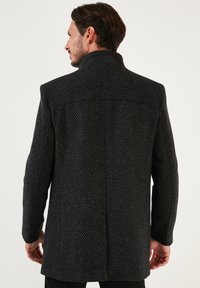 Abrigo texturizado oscuro con un cuello alto, que presenta un patrón sutil y un corte recto. La tela parece duradera y es de color gris oscuro.