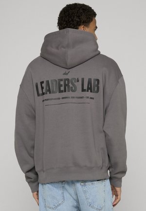 Man wearing gray hoodie with hood up, black text "LEADERS' LAB" on back, paired with light blue jeans, facing away.