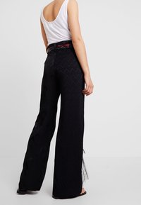 Pantalon noir à jambes larges avec un motif en zigzag texturé, orné de franges à l'ourlet et d'une ceinture à motifs aux couleurs contrastantes.