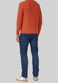 Man draagt donkerblauwe jeans, witte sneakers en een oranje shirt met lange mouwen, staand tegen een effen grijze achtergrond.