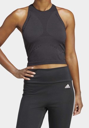 Vrouw draagt een zwarte, mouwloze, aansluitende cropped top met perforaties en zwarte Adidas legging met witte strepen aan de zijkanten en een logo op de taille.