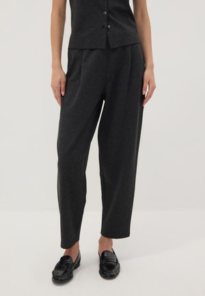 Donna che indossa pantaloni larghi grigio scuro, mocassini neri e un top abbottonato grigio scuro abbinato, in piedi con le mani rilassate lungo i fianchi.
