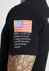 Black t-shirt featuring a stitched US flag patch and white text. Text reads: "NASA's mission is to pioneer the future in space exploration..."