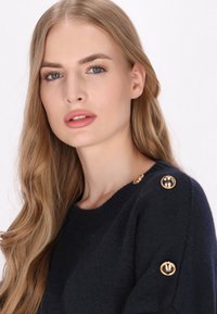Pulover bleumarin dintr-un material moale, cu nasturi aurii rotunzi la umeri, având un gât clasic și o croială lejeră.