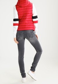 Röd pufferväst med huva, bärs över en vit sweatshirt med svarta och röda ränder. Matchas med slim-fit gråjeans och vita sneakers.