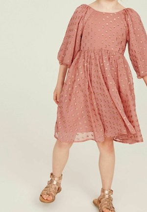 Robe rose à pois avec des manches bouffantes, texture superposée et accents dorés. Associée à des sandales gladiateurs métalliques.