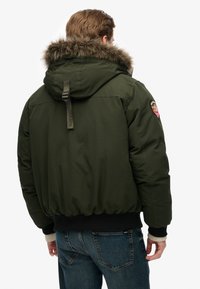 Doudoune vert olive avec capuche en fourrure, poignets et taille côtelés, comportant un patch logo sur la manche gauche.