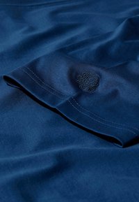 Jūras pudelē zils kokvilnas audums ar gludu faktūru; ar mazu melnu izšūtu logotipu uz piedurknes un smalkiem šuvēm.