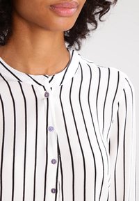 Blouse blanche à rayures verticales noires, dotée d'un col mandarin, de boutons à l'avant et de boutons violets brillants. Tissu doux et texturé.