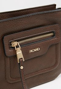 Ruskeasta nahasta valmistettu olkalaukku, jossa on teksturoitu pinta, kultainen vetoketju ja etutasku, jonka reunus on musta. Brändin nimi "PICARD" on kohokuvioitu.