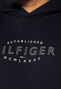 Felpa nera con cappuccio caratterizzata dalla scritta "ESTABLISHED ILFIGER MCMLXXXV" in bianco e rosso, con una texture in cotone morbido. Design con cappuccio e vestibilità casual.