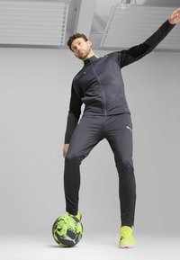 Athletisk outfit i mørkegrå, bestående af en zip-up jakke med sorte ærmer og grønne detaljer, tætsiddende bukser og neongrøn fodboldsko, stående på en mønstret bold.