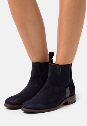Bottines en daim bleu marine avec des panneaux latéraux élastiques, bouts ronds et petit talon marron ; présente un accent logo doré sur le côté.