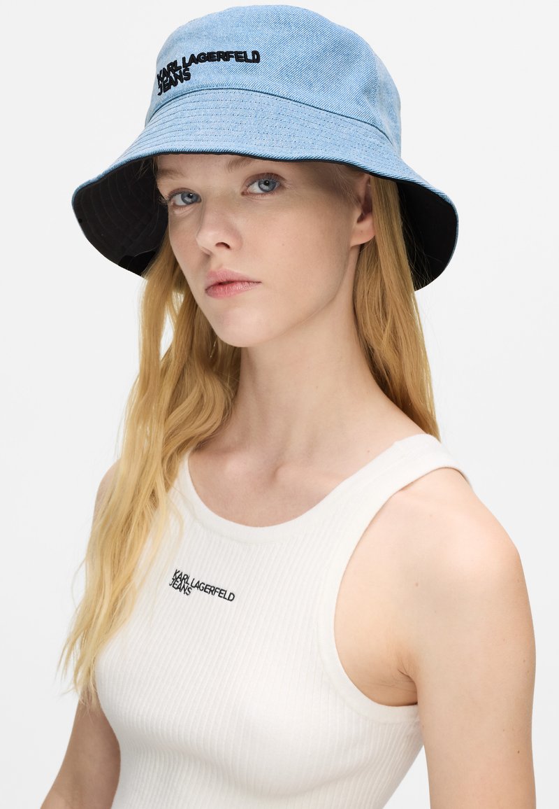 Cappello a secchi light blue in denim con interno nero, dotato di logo ricamato "Karl Lagerfeld Jeans". Modello che indossa una canotta bianca a coste.