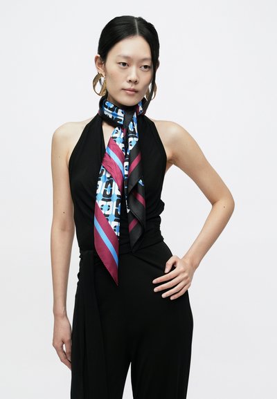 Svilena ruta z modro in črno geometrijsko vzorcem, poudarjena z izrazitimi diagonalnimi črtami v roza in modri barvi, nošena okoli vratu.