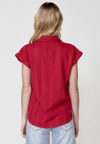 Rode kortemouwshirt met een gestructureerd, gestreept patroon; heeft een kraag en een afgeronde zoom. Gecombineerd met lichtblauwe denim jeans.